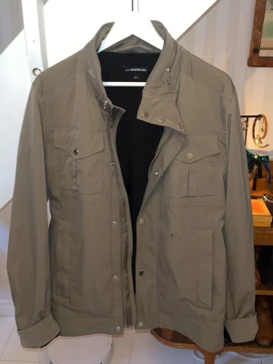 Olivgrön fieldjacket från J.Lindeberg - Stilren olivgrön fieldjacket från J.Lindeberg med hög krage, dragkedja och tryckknappar. Fyra fickor framtill med lock och knappar samt justerbara detaljer bak. Tillverkad i ett lätt och tåligt material, perfekt för blåsiga dagar. Den är lite smutsig men går att tvätta bort. Priset är ej hugget i sten så tveka inte på att ställa frågor😊mvh Oscar 😊🙌