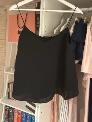 Svart satin linnetopp från Stradivarius - Säljer en svart satin linnetopp från Stradivarius med tunna axelband och v-ringad hals. Toppen har en enkel och stilren design, perfekt att matcha med olika outfits. Materialet känns lätt och luftigt.