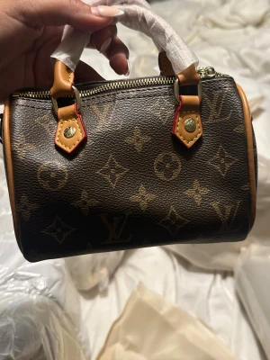 monogram handväska mini - Säljer en liten och ikonisk handväska i klassiskt brunt monogrammönster. Väskan har två korta handtag i ljust skinn, guldfärgade metalldetaljer och dragkedja upptill. Perfekt accessoar för dig som gillar lyxig och tidlös design.