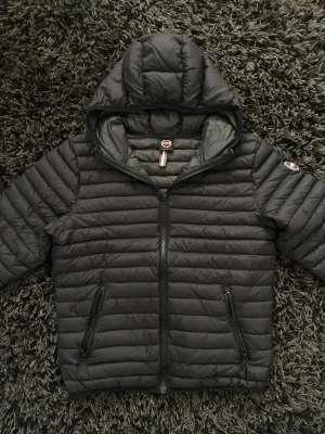 Svart dunjacka från Colmar - Snygg svart dunjacka från Colmar med huva och två dragkedjeförsedda fickor. Jackan har quiltad design och Colmar-logga på ärmen. Perfekt för kalla vinterdagar och riktigt skön att bära. Materialet är lätt och smidigt.