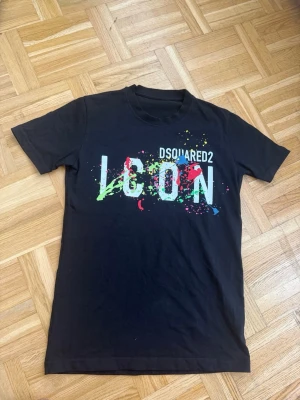 Svart Dsquared2 ICON t-shirt - Snygg svart t-shirt från Dsquared2 med stort vitt 'ICON'-tryck och färgglada färgstänk över bröstet. Klassisk rund hals och korta ärmar. Perfekt för dig som vill sticka ut med en trendig och ungdomlig look. Storlek S helt ny och oanvänd.