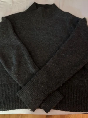 Mörkgrå stickad polotröja - Mysig mörkgrå stickad polotröja med ribbade muddar vid ärmslut och nederkant. Tröjan har en hög hals och är tillverkad i ett mjukt, tjockt material som passar perfekt för kyliga dagar. Enkel och stilren design som är lätt att matcha. Storlek S