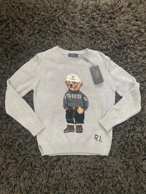 Grå stickad tröja med björnmotiv Polo - Grå stickad tröja från Polo Ralph Lauren med ikoniskt björnmotiv på bröstet. Tröjan har rund hals, ribbade muddar och RL-broderi nedtill. Perfekt för dig som gillar klassisk men lekfull stil.