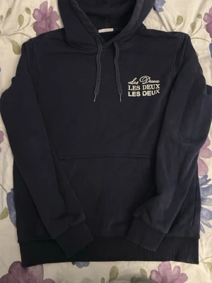  hoodie  Les Deux - Snygg mörkblå hoodie från Les Deux med vit broderad logga på bröstet. Klassisk känguruficka framtill, ribbade muddar och justerbar huva med dragsko. Perfekt för en avslappnad och trendig stil.