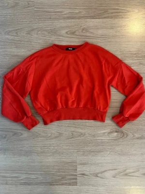 Röd croppad sweatshirt från Bik Bok - Säljer en röd croppad sweatshirt från Bik Bok med rund halsringning och breda muddar vid ärmslut och midja. Tröjan har en enkel och clean design utan tryck eller detaljer, perfekt för en avslappnad och trendig look.
