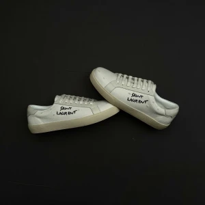 Vita Saint Laurent sneakers - Snygga vita sneakers från Saint Laurent med handskriven logga på sidan. Skorna har klassisk låg profil, vita snören och en stilren gummisula. Tillverkade i canvas med detaljer i läder för en clean och modern look.