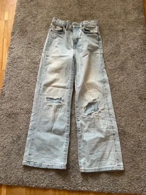 Ljusblåa vida jeans med hål - Säljer ett par ljusblå jeans med raka, vida ben och slitna hål på båda knäna. Jeansen har klassisk femficksdesign och är tillverkade i denim. Perfekta för en avslappnad och trendig stil.