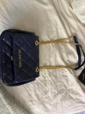 Mörkblå axelväska från Valentino - Snygg mörkblå axelväska från Valentino med quiltat mönster och guldfärgade kedjor. Väskan har en klassisk rektangulär form och lock med logga i guld. Inga defekter och använd fåtal gånger. Pris kan diskuteras 
