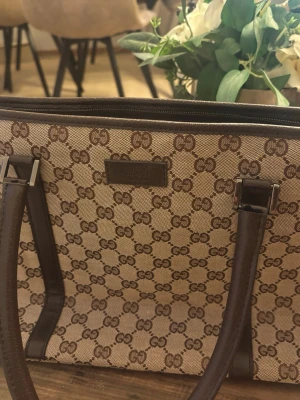Beige och brun handväska från Gucci - Snygg handväska från Gucci i beige canvas med klassiskt GG-mönster och mörkbruna läderdetaljer. Väskan har två långa handtag och silverfärgade metalldetaljer. Perfekt för dig som gillar tidlös och lyxig design.