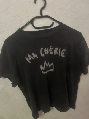 Svart t-shirt med tryck 'Ma Chérie' - Cool svart t-shirt med vitt tryck där det står 'Ma Chérie' och en krona på bröstet. Klassisk passform och rund halsringning, perfekt till jeans eller kjol. T-shirten har korta ärmar och är i mjukt material.
