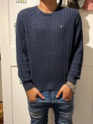 Gant stickad tröja  - Mörkblå kabelstickad tröja med rund halsringning och ribbade muddar vid ärmslut och nederkant. Tröjan har ett diskret broderat märke på bröstet och är tillverkad i ett mjukt stickat material. Perfekt för en avslappnad och stilren look.