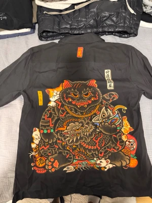 Svart skjorta med drak- och kattmotiv - Unik svart skjorta med kort ärm och knappar framtill. Stora färgglada tryck på ryggen med katter och asiatiska motiv samt en drake och röd cirkel på framsidan. Skjortan har en avslappnad passform och är tillverkad i ett mjukt tyg, perfekt för dig som gillar statement-plagg. Storlek S passar M