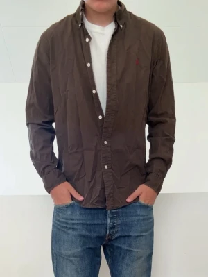 Mörkbrun Ralph Lauren skjorta - Snygg brun skjorta med långa ärmar och klassisk krage. Skjortan har vita knappar framtill och en liten broderad detalj på bröstet. Perfekt att bära öppen över en t-shirt eller stängd för en mer uppklädd look