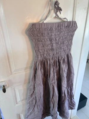 Ljuslila  klänning från kay/day - Säljer en ljuslila klänning med smock upptill och voluminös kjol. Klänningen har knytband i nacken och är axelbandslös, perfekt för varma dagar. Tillverkad i ett mjukt och lätt tyg som ger en avslappnad look.