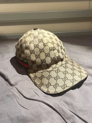 Äkta beige Gucci keps - Säljer en äkta beige keps från Gucci med det klassiska GG monogrammet över hela kepsen. På sidan finns en grön och röd rand. Kepsen har en justerbar rem bak. Perfekt under sommaren, dm för fler frågor💯