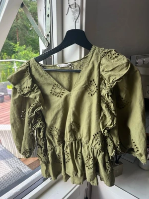 Olivgrön blus med volanger - Snygg olivgrön blus med v-ringning och broderade håldetaljer. Blusen har volanger längs axlarna och ärmarna samt en lös, flowy passform. Perfekt för dig som gillar en bohemisk och trendig stil.