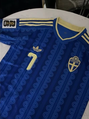 Blå Sverige fotbollströja Adidas - Säljer en blå fotbollströja från Adidas med gula detaljer, nummer 7 på bröstet och Sveriges landslagsemblem. Tröjan har klassiska Adidas-ränder på axlarna och ett mönster i tyget. Perfekt för dig som vill stötta Sverige på match eller träning.