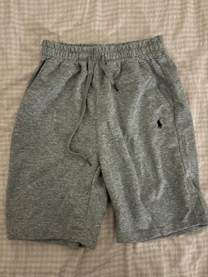 Grå mjukisshorts från Polo Ralph Lauren - Gråa mjukisshorts från Polo Ralph Lauren med elastisk midja och dragsko. Klassisk logga broderad på vänster ben. Tillverkade i mjukt bomullsmaterial och har två sidofickor. Perfekta för en avslappnad och sportig stil.