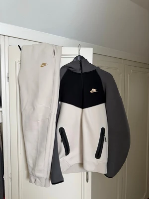 Nike tech fleece komplett  - Nike komplett i färgerna svart, grå och beige med dragkedja framtill och två fickor med svarta detaljer. Guldigt Nike-märke på bröstet och huva för extra stilpoäng. Perfekt för chill dagar eller när du vill ha en sportig look.