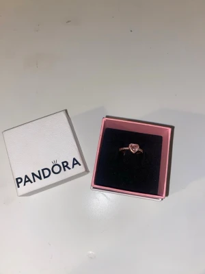 Roséguldsring med hjärta från Pandora - Supersöt ring från Pandora i roséguld med ett gnistrande hjärta i mitten, omgivet av små klara stenar. Ringen har en tunn, elegant ringband med stenar även på sidorna. Perfekt för dig som gillar romantiska detaljer och vill ha något som sticker ut. Och den kommer även med kartongen som man kan se på bilden.