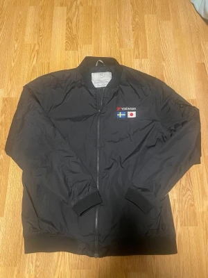 Svart bomberjacka med YOKOHAMA-tryck - Svart bomberjacka från Nimbus med dragkedja framtill och muddar vid ärmslut och midja. På bröstet finns broderat YOKOHAMA-logga samt svenska och japanska flaggan. Klassisk passform och stilren look, perfekt för dig som gillar streetwear.