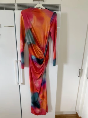 Maxiklänning med fina färger - Säljer en långklänning i färgstarkt mönster med inslag av orange, rosa, blått och svart. Klänningen har lång ärm och en snygg drapering i sidan som ger en cool effekt. Materialet känns stretchigt och följsamt, perfekt för dig som vill sticka ut.  Använd endast 1 gång perfekt skick.