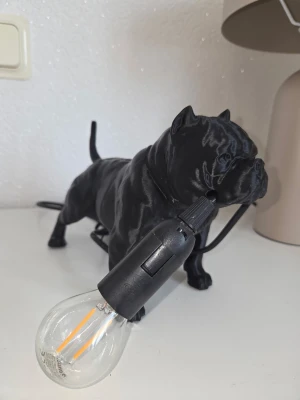 Bordslampa Pitbull  - Unik bordslampa formad som en svart Pitbull i plast. Hunden håller en glödlampa i munnen, vilket ger en cool och modern känsla till rummet. Perfekt som dekoration eller mysbelysning i hemmet.
