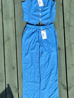 Blått tvådelat set med väst och byxa - Snyggt blått set med en ärmlös väst med knappar framtill och matchande vida byxor med resår i midjan. Setet har en modern look och är gjort i ett lätt tyg som passar perfekt till sommaren. Västen har en figurnära passform och byxorna har fickor.