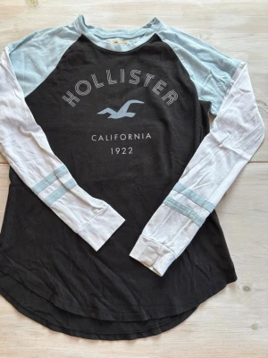 Långärmad tröja från Hollister - Långärmad tröja från Hollister med svart kropp, ljusblå axelparti och vita ärmar med ljusblå ränder. Stor logga och text framtill. Tillverkad i mjuk bomull för en skön känsla.