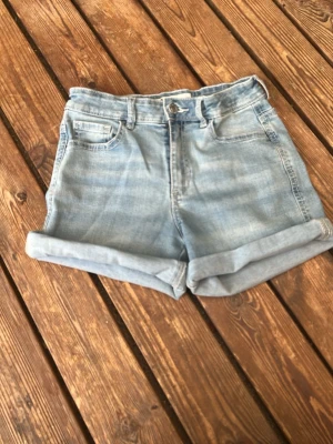 Ljusblå denimshorts med fickor - Säljer ett par ljusblå denimshorts med klassisk fyrfickig och knappgylf. Shortsen har en rak passform och kan vikas upp för en mer avslappnad look. Perfekta till sommaren och lätta att matcha med olika stilar. Har en liten fläck framvid men syns inte så tydligt