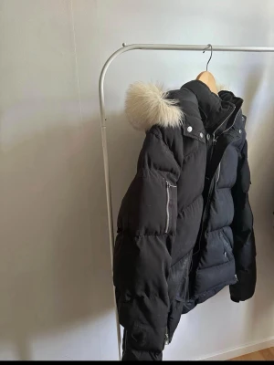 Moose Knuckles jacka - Säljer en svart pufferjacka med stor huva och fuskpälsdetalj. Jackan har dragkedjor, tryckknappar och en patch på ärmen. Flera fickor och en oversized look gör den både snygg och praktisk. Perfekt för kalla vinterdagar.