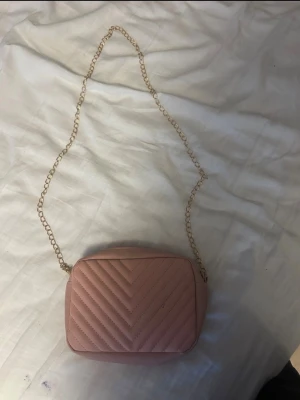 Rosa quiltad axelväska med kedja - Säljer en snygg rosa axelväska med quiltat mönster och guldfärgad kedjerem. Väskan har en rektangulär form och är tillverkad i ett slätt material som liknar konstläder. Perfekt för dig som vill ha en trendig och stilren accessoar.