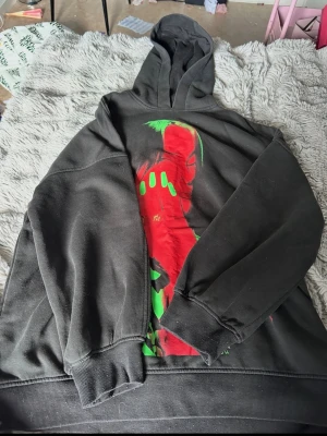 Svart hoodie med neontryck - Cool svart hoodie med stor färgstark print på framsidan i rött och grönt som föreställer ett ansikte och hand med neonfärgade detaljer. Hoodien har huva och ribbade muddar vid ärmar och nederkant. Perfekt för dig som gillar streetwear och unika plagg.