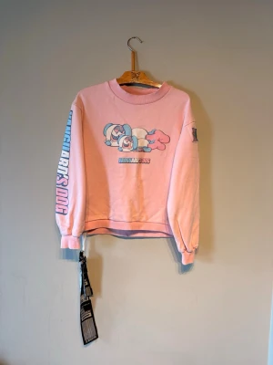 Rosa sweatshirt med Vanguard's Dog-tryck - Säljer en rosa sweatshirt med tryck av två tecknade hundar och texten 'Vanguard's Dog' på bröstet. Tröjan har ribbade muddar, rund hals och tryck på ena ärmen. Perfekt för dig som gillar streetwear och unika prints.