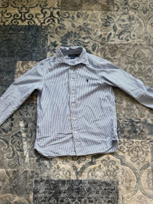 Vit och blå randig Ralph Lauren skjorta storlek L barnstorlek - Klassisk skjorta med smala blå och vita rände från Ralph Lauren, långärmad och med knappar framtill. Skjortan har en krage. Perfekt för en clean och stilren look.