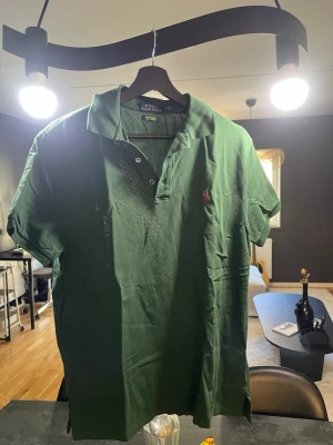 Grön pikétröja från Polo Ralph Lauren - Äkta grön Ralph Lauren Polo tröja  Custom Slim Fit Stretch Mesh. Med röd logga på.  Köpt i Singapore, säljer min killes tröja som han inte använder längre och har använts 2 gånger bara.   Är i bra skick fortfarande, inga skador, hål eller fläckar på tröjan.   Storlek: Medium Axel längd: 43cm Arm längd: 17cm Längd: 67cm Bredd: 49cm