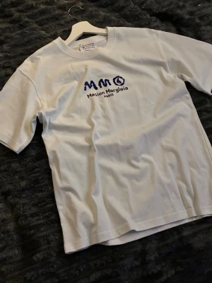Vit Maison Margiela t-shirt - Vit t-shirt från Maison Margiela med blå logga och text på bröstet. Endast använts 2 gånger, säljs pga för liten.