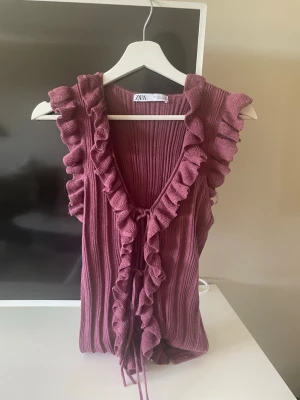 Vinröd ribbad linnetopp med volang - Supersöt vinröd linnetopp från ZARA med ribbad struktur och volangkant längs ärmhål och framkant. Toppen har knyt framtill och är ärmlös, perfekt för en trendig och feminin look. Storlek 40 men känns mindre