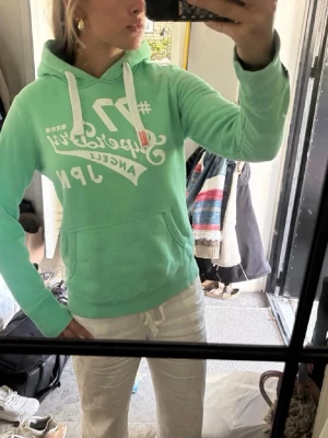 Mintgrön hoodie från Superdry - Snygg mintgrön hoodie från Superdry med vit tryckt logga och text på bröstet. Hoodien har vita dragsnören, känguruficka och ribbade muddar. Perfekt för en avslappnad och cool stil (hoodien passar xs-s men eftersomatt det är från super dry har de väldigt små storlekar) .