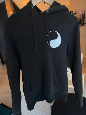Svart hoodie från Stussy med tryck - Svart hoodie från Stussy med en stor ficka framtill och justerbar huva. På bröstet finns ett vitt grafiskt tryck. Hoodien har ribbade muddar vid ärmar och nederkant samt klassiska dragsnören i huvan. Perfekt för en avslappnad streetwear-look.