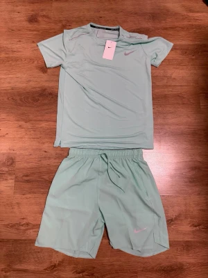  Nike Set - Snyggt mintgrönt träningsset från Nike med t-shirt och shorts. Båda plaggen har en enkel design med Nike-logga i ljusrosa. T-shirten har rund hals och korta ärmar, shortsen har resårmidja med dragsko och sidofickor. Perfekt för gym eller löpning.Står storlek L men passar S