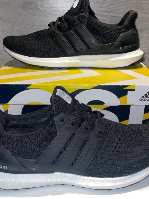 Svarta Adidas Ultra Boost sneakers - Svarta Adidas Ultra Boost sneakers med stickad ovandel i mesh och klassiska tre ränder på sidan. Skorna har en vit Boost-mellansula för extra komfort och svart yttersula. Snygg och sportig design med plastdetaljer vid snörningen.