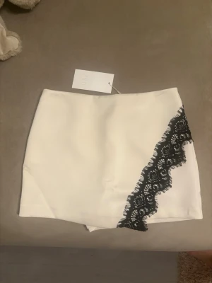Vit skort med svart spetsdetalj - Supersnygg vit skort med en unik svart spetsdetalj på framsidan. Skorten har en rak och stilren passform med dragkedja bak. Perfekt för dig som vill ha kjol-looken men ändå känna dig bekväm. Materialet känns mjukt och följsamt.