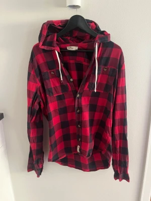 Röd och svart rutig hoodie-skjorta från Hollister - Säljer en röd och svart rutig skjorta med huva och knappar framtill. Skjortan har två bröstfickor med knapp och vita snören i huvan. Perfekt för en avslappnad och cool stil. Storlek L men skulle säga att den passar M lite bättre. 