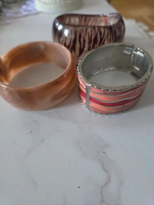 Set med tre breda armband - Säljer ett set med tre breda armband i olika färger och material. Ett armband är i brunmelerad plast, ett i lila med vitt mönster och ett i metall med rosa och röda ränder. Perfekta för att ge din outfit en färgklick och unik touch.