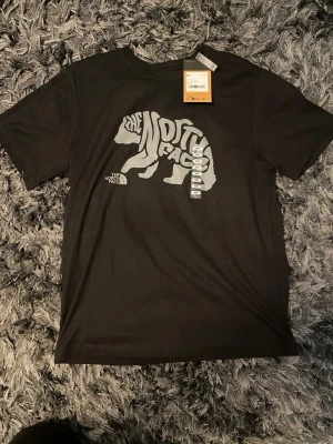 Svart t-shirt The North Face  - Snygg svart t-shirt från The North Face med stor björnprint och logga i vitt på bröstet. Klassisk passform och rund halsringning. Tillverkad i mjuk bomull som känns skön mot huden. Perfekt för dig som gillar streetwear och outdoor-vibe.