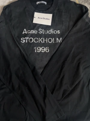 Svart Acne Studios långarmad t-shirt med tryck - Cool svart t-shirt från Acne Studios med vit text 'Acne Studios STOCKHOLM 1996' på bröstet. Klassisk rund halsringning och mjukt bomullsmaterial. Perfekt för dig som gillar stilrena och trendiga plagg med statement-tryck. Pris kan diskuteras 