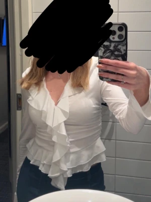 Vit omlottblus med volangdetaljer - Säljer en vit omlottblus med långa ärmar och snygga volangdetaljer längs urringningen och framtill. Blusen har en v-ringad skärning och markerad midja som ger en feminin siluett. Perfekt att styla med jeans eller kjol för en trendig look.