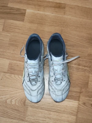Puma king pro - Säljer ett par vita fotbollsskor med svarta detaljer och snörning. Skorna har dobbar för spel på gräs och en sportig design med diskreta linjer på sidorna. Perfekta för dig som vill ha stilrena och funktionella skor på planen.