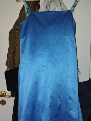 Blå axelbandslös klänning - Säljer en enkel och stilren blå klänning med tunna axelband. Klänningen har en rak passform och är tillverkad i ett glansigt material som ger en snygg look. Perfekt för dig som gillar minimalistisk stil.